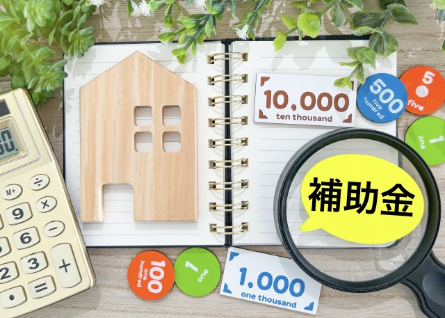 補助金等の情報お知らせ記事に使用した補助金が虫眼鏡でアップし、周りに家や電卓が並んだ画像