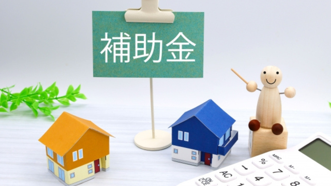 補助金等の情報お知らせ記事に使用した補助金と書かれた掲示板を人形が棒で刺し、周りに家の模型があるの画像